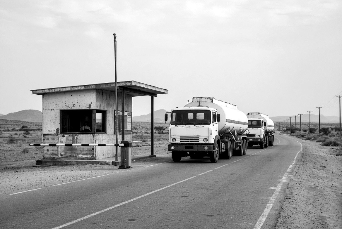 Two tankers queued on a road at a checkpoint barrier beside a small booth.