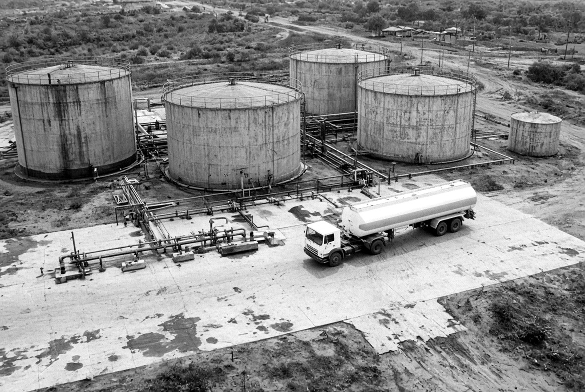 Aerial view of five cylindrical storage tanks and connecting pipework, with a road tanker parked on a concrete apron.