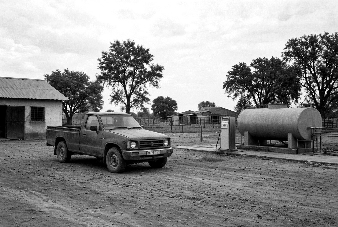 A pickup truck parked on a dirt yard beside a fuel pump and a horizontal tank.