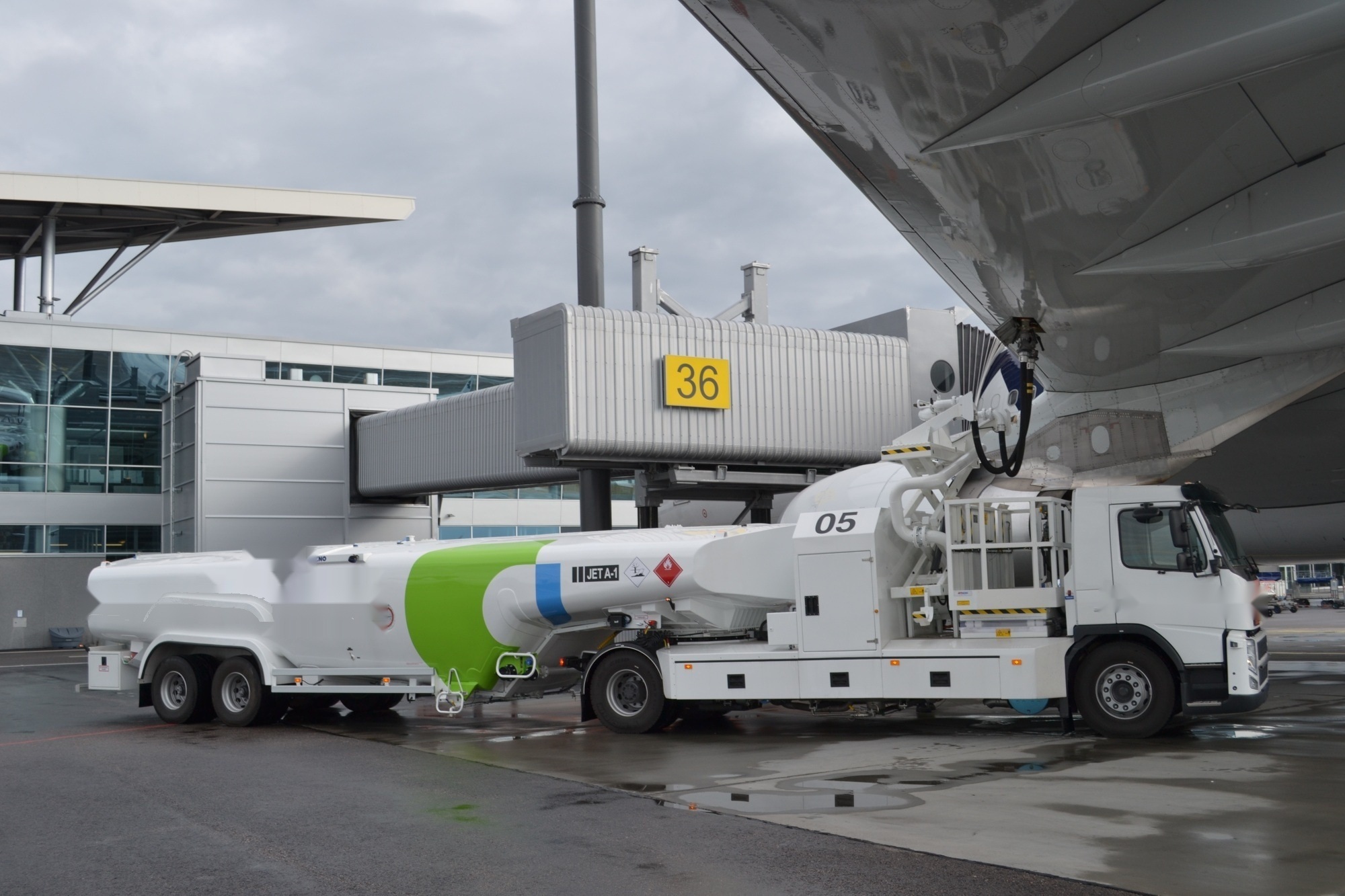 A refuelling tanker parked beneath an aircraft wing at an airport stand, with a hose connected to the wing and a boarding bridge behind.