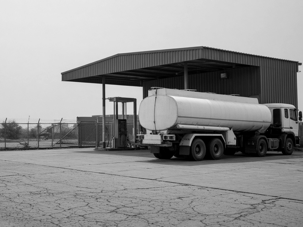 A road tanker parked under a steel loading canopy on a concrete apron, beside a dispensing point.